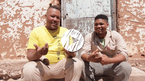 Two men sitting on the ground outside a weathered building with peeling paint and a wooden door, smiling and engaging with the camera.